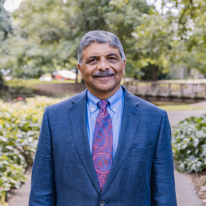 Headshot image of president Kaushik Kotecha smiling, wearing a blue suit and pink tie. Background is a park.